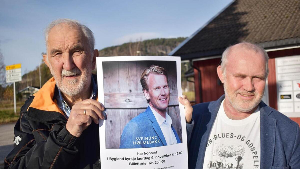 Harry Andersen (t.v.) og Olav Nilsen med plakat som fortel om Sveinung Hølmebakk sin konsert i Bygland kyrkje førstkomande laurdag.